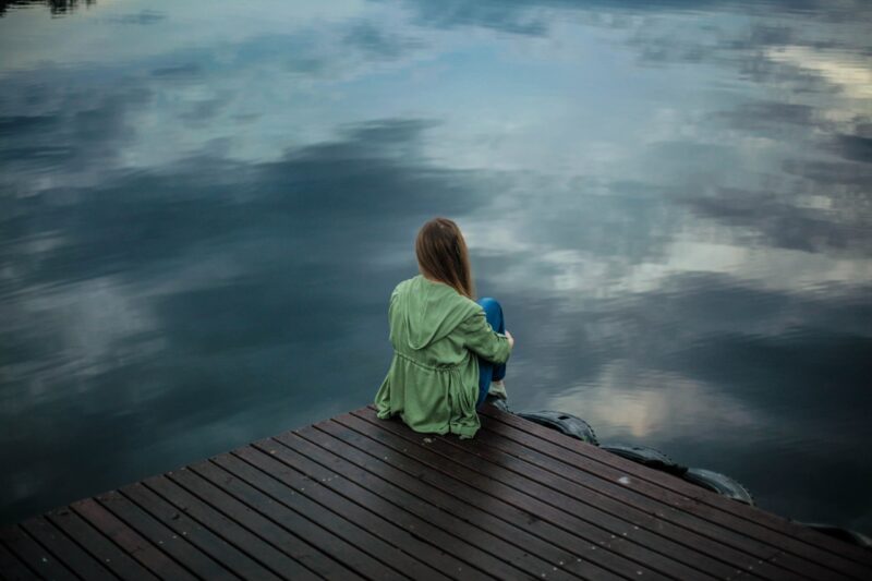 Frau sitzt alleine auf einem Steg am Wasser